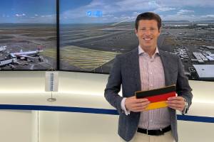 Marcel Haar stehend vor Bildschirmen, die einen Flughafen zeigen; er hält eine deutsche Fahne in der Hand